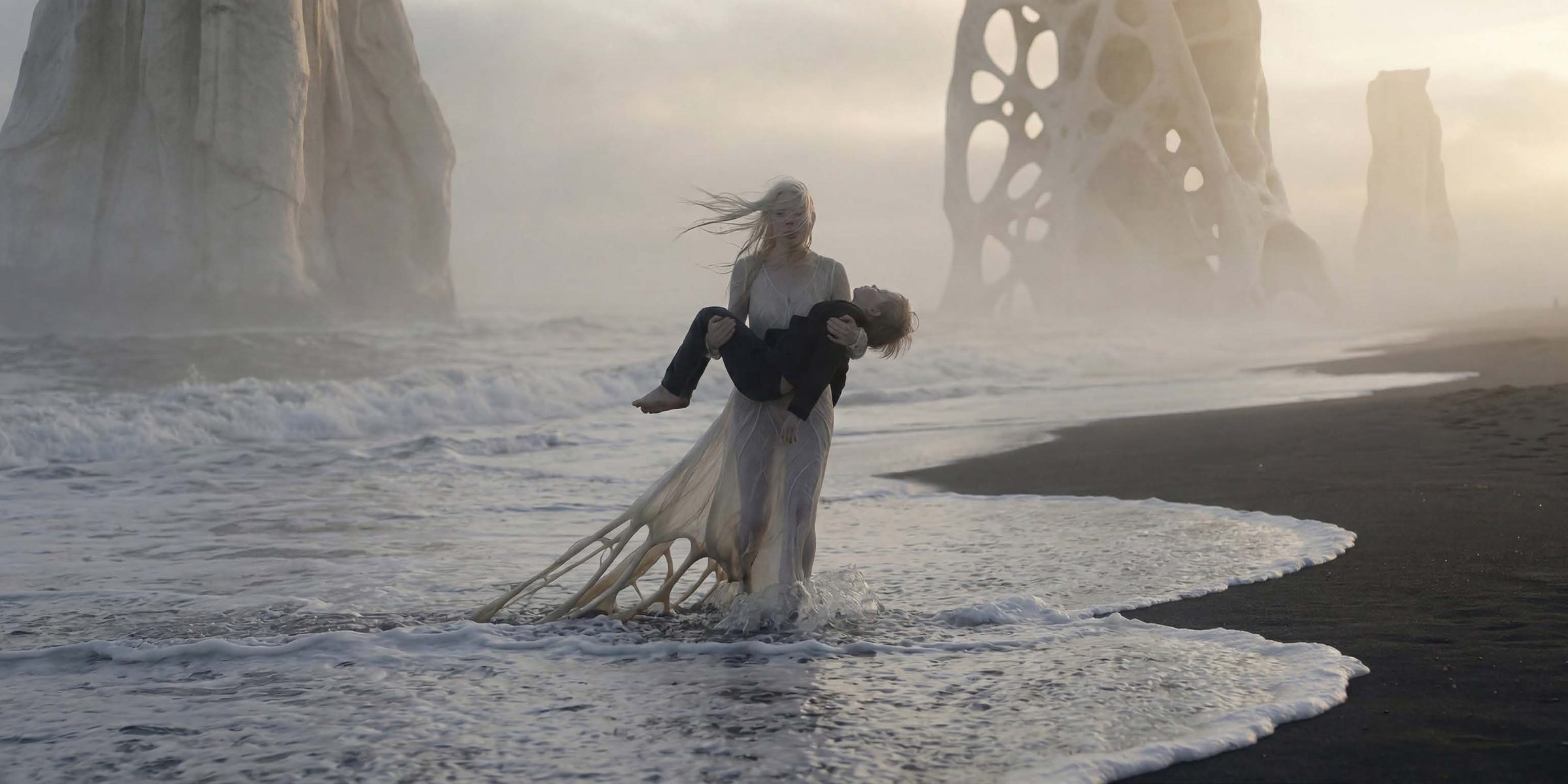 A woman with long pale blonde hair blowing sideways in the wind stands shin-deep in breaking surf on a black volcanic sand beach, carrying an unconscious man dressed in black in her arms. His head is thrown back over her