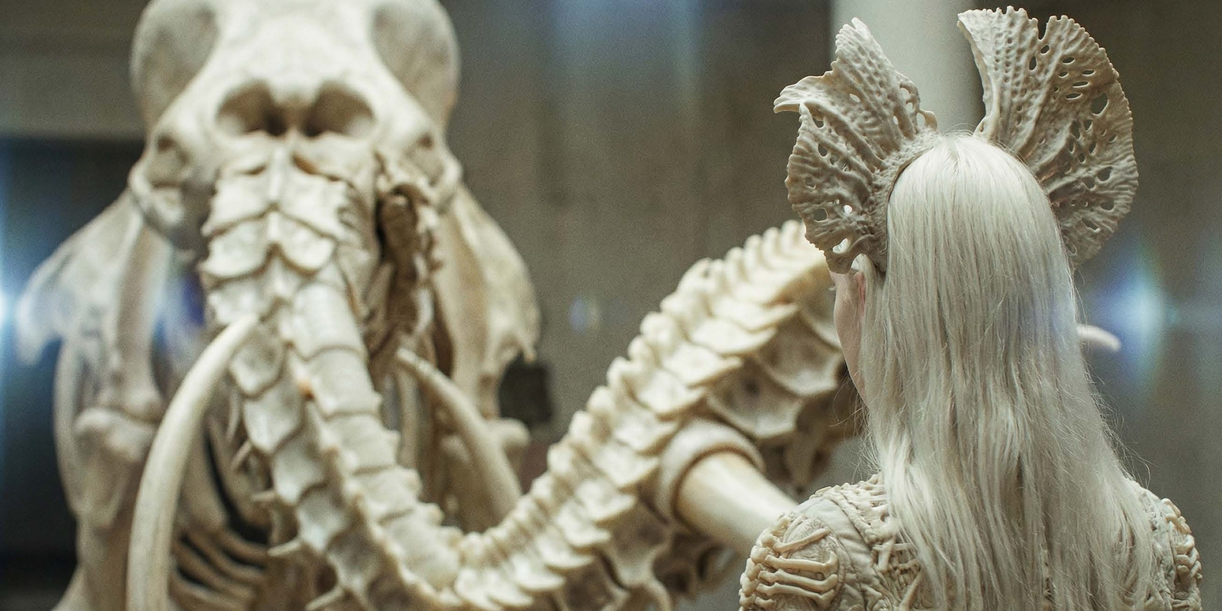 A figure with long platinum-white hair, seen from behind and wearing an intricately bone-carved gown and a large perforated fan-like bone headdress, stands at right facing a colossal ivory-white creature skull with a smo